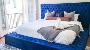 Een slaapkamer met een blauw, gestoffeerd hoofdbord, witte beddengoed en een zwaan-vormig kussen.