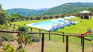 Ein Swimmingpool mit Liegestühlen und Sonnenschirmen, umgeben von einem eingezäunten Garten mit Blick auf grüne Hügel.