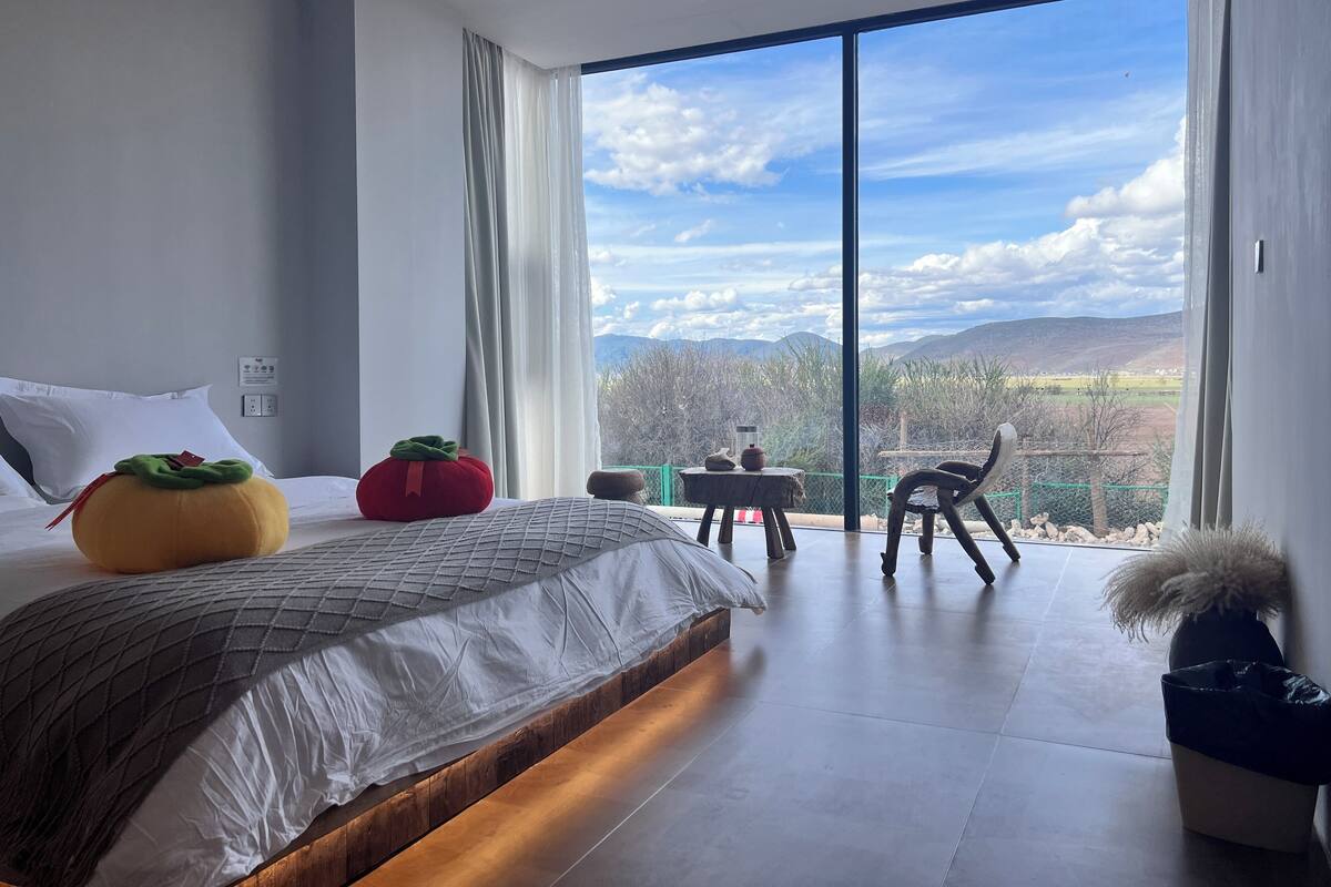 Quarto moderno com uma cama grande, vista para montanhas e varanda com cadeira e mesa.