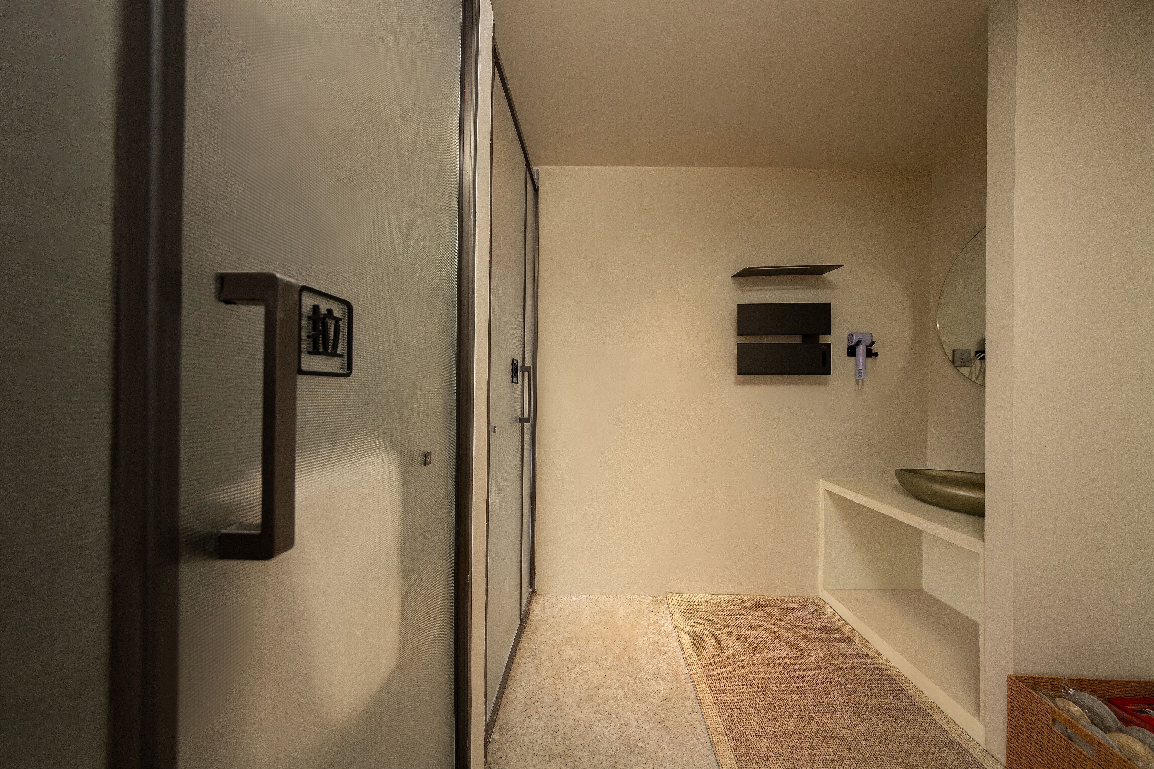 Un pasillo estrecho con una puerta a la izquierda, una pared blanca y un baño con lavamanos y espejo a la derecha.