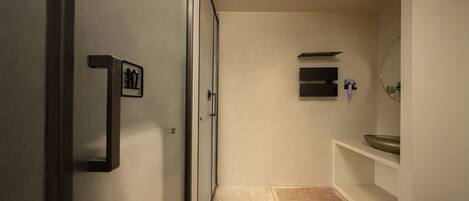 Un pasillo estrecho con una puerta a la izquierda, una pared blanca y un baño con lavamanos y espejo a la derecha.