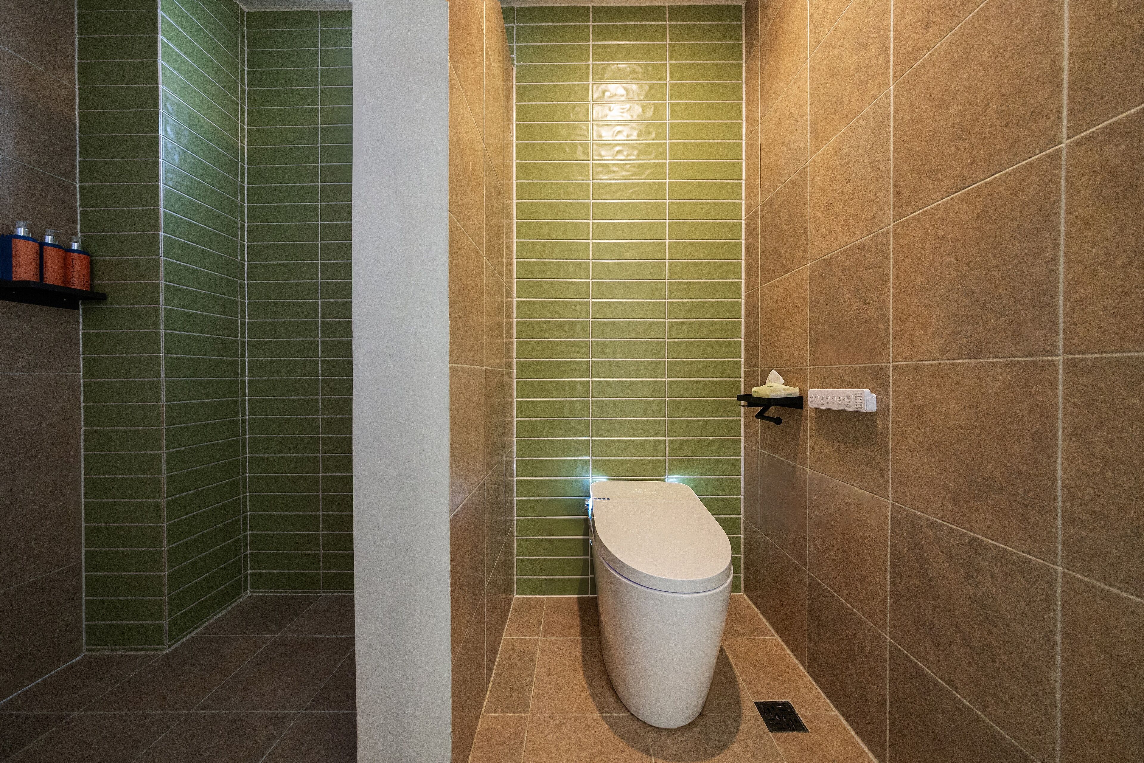 Un baño con baldosas verdes y beige, un inodoro blanco y un estante con artículos de aseo.