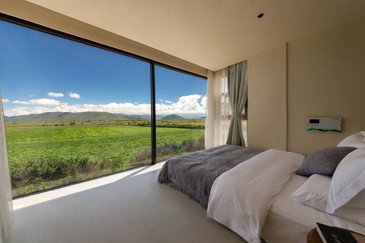 Quarto com uma grande janela que oferece vista para um campo verde e montanhas ao longe.