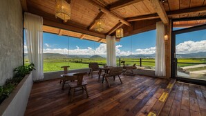 A wooden deck with chairs and tables, large windows with curtains, and a view of a green landscape. - LOST VILLA-ff (Deqin)