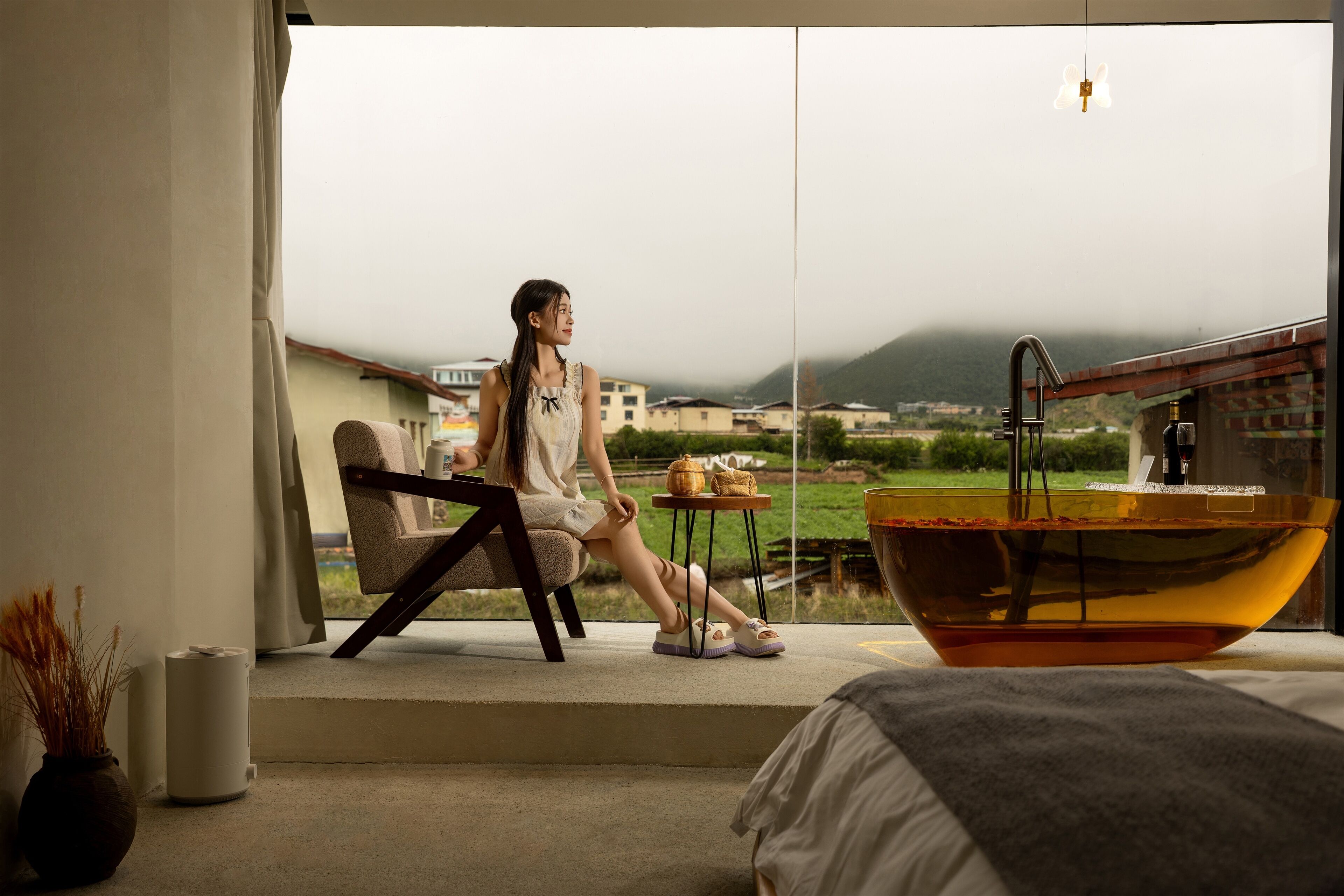 A room with a large window, a bed, a chair, a table with a teapot, and a large bowl.