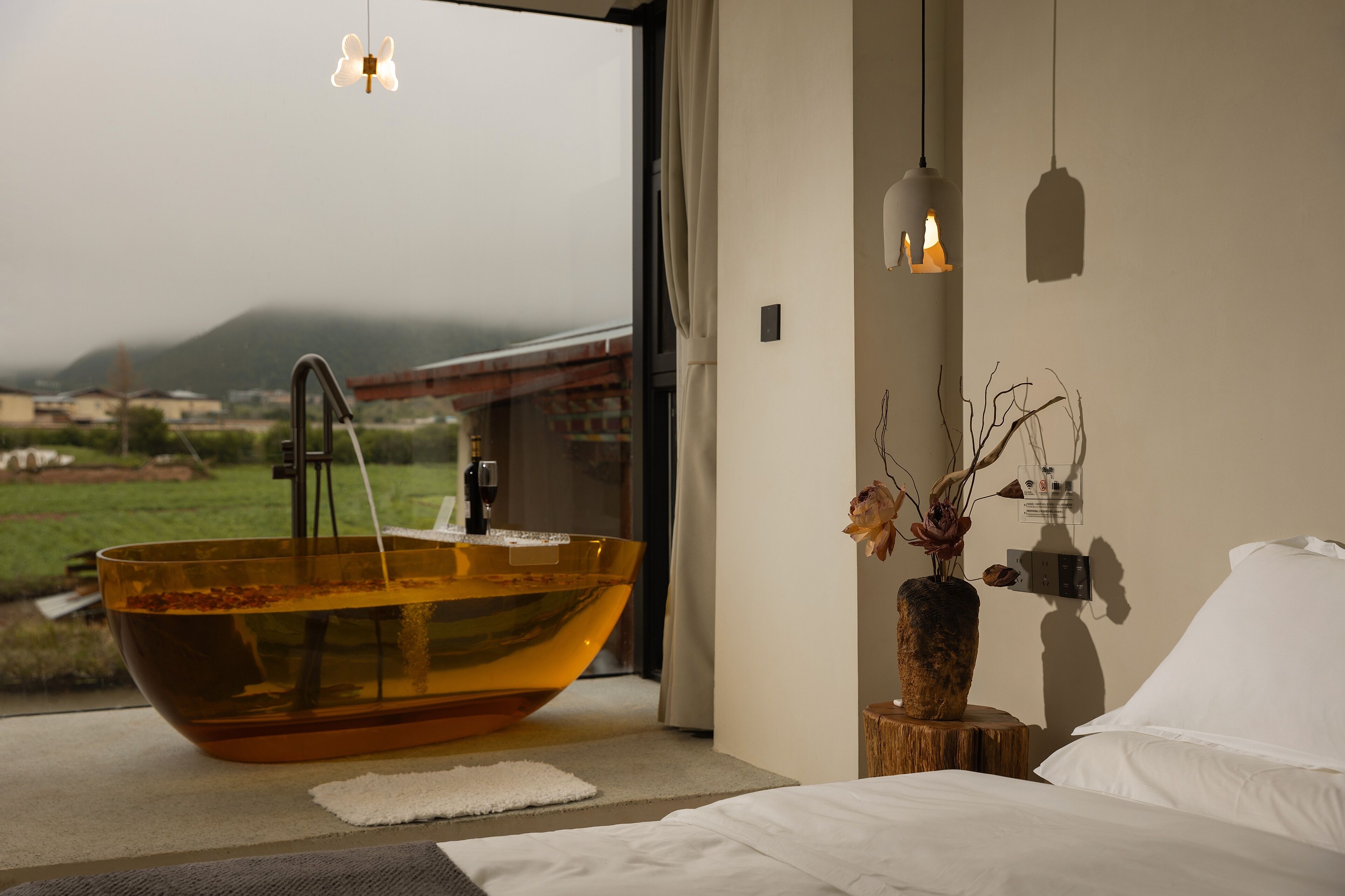 Un baño moderno con una bañera de vidrio grande, un taburete de madera con un florero, y vistas a un paisaje verde.