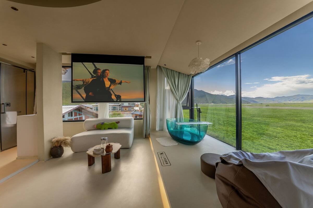 Sala de estar moderna com uma televisão grande, uma tigela de vidro e vista para um campo verde e montanhas.