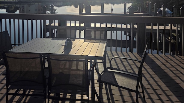 Una terraza de madera con mesas y sillas, con vistas a un cuerpo de agua tranquilo y edificios al fondo.