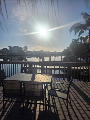 A wooden deck with tables and chairs, overlooking a calm body of water and buildings in the background.