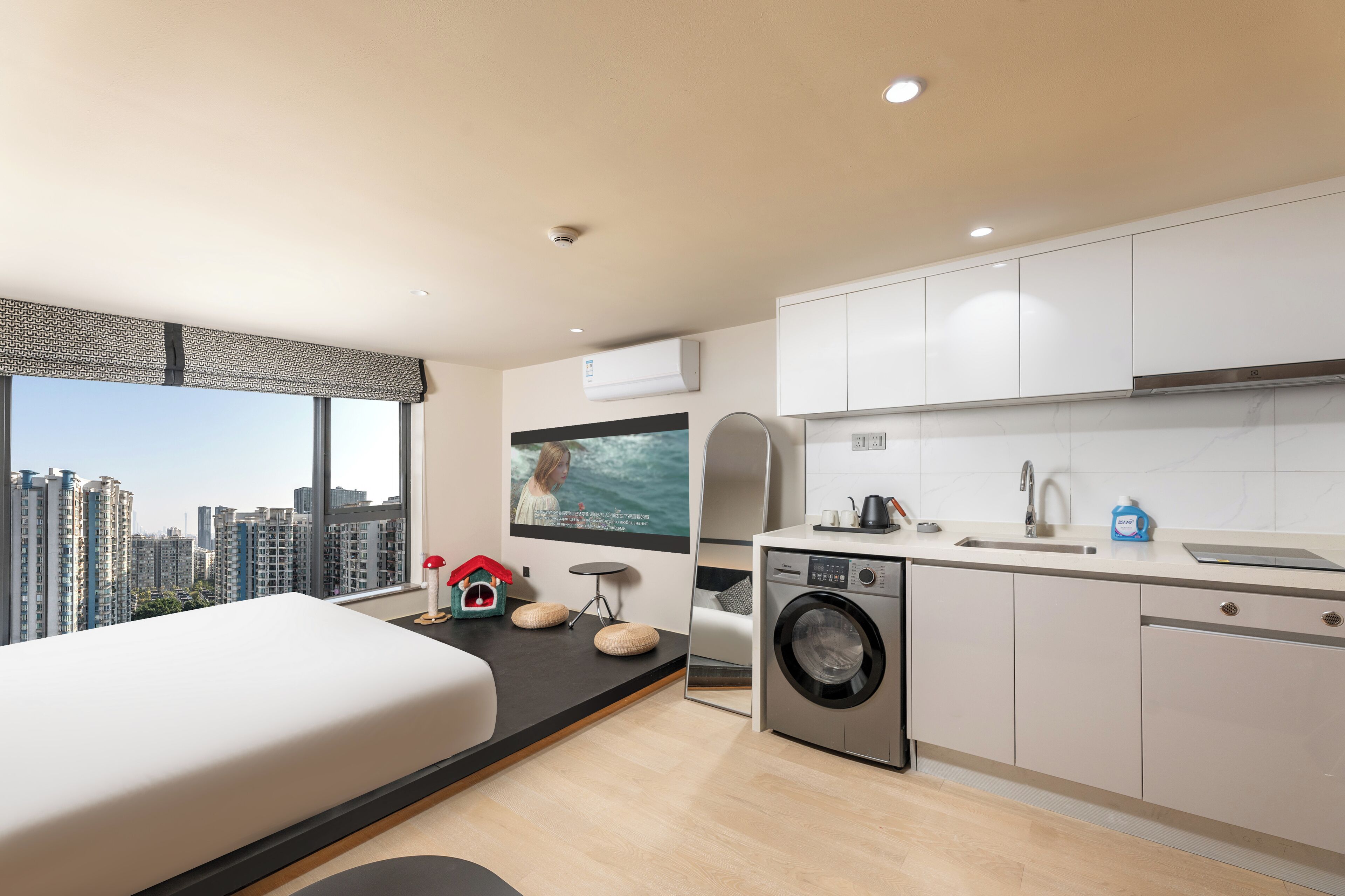 A modern hotel room with a bed, a washing machine, a sink, and a large window offering a city view.