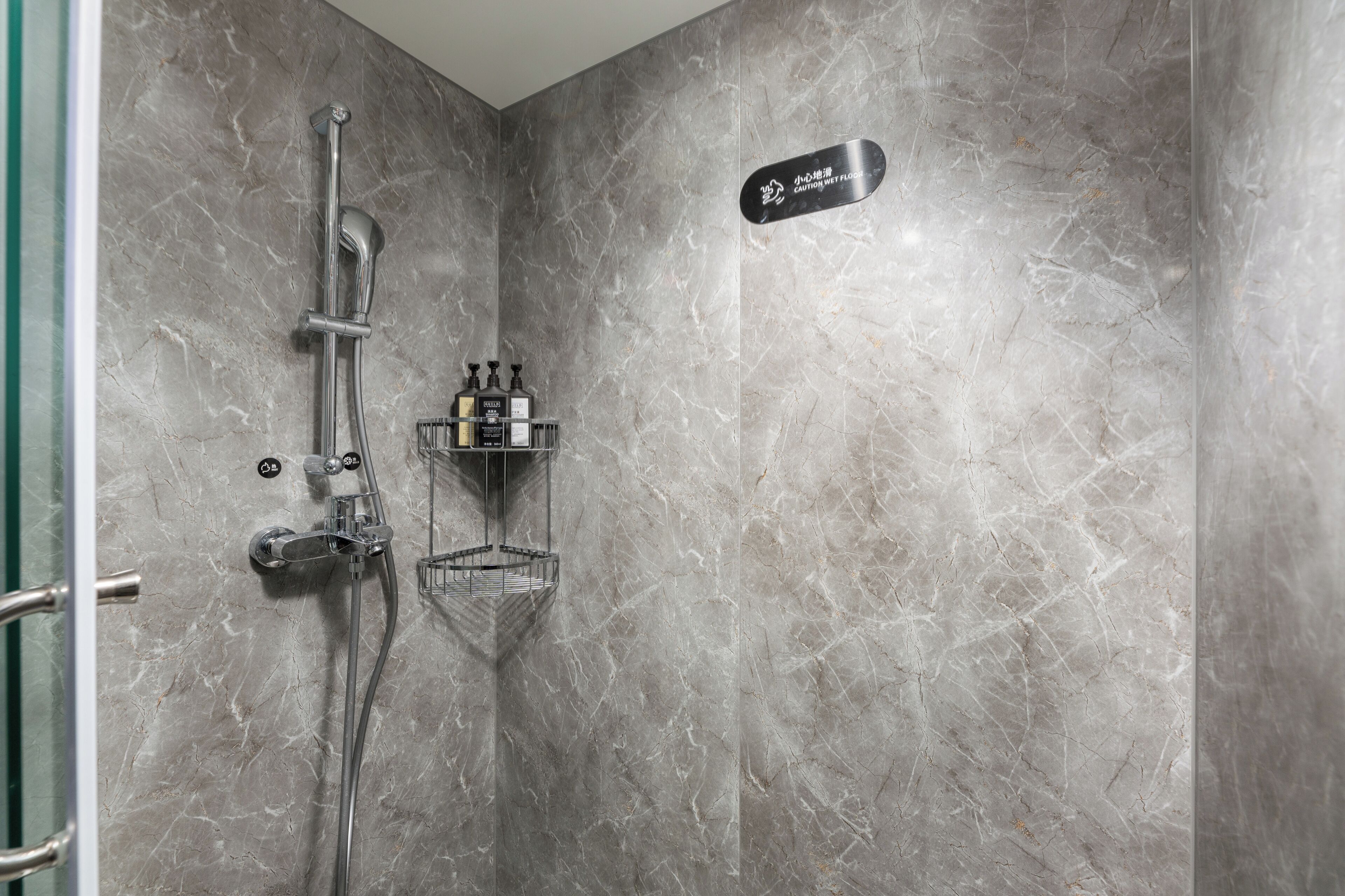 Un baño con ducha, un cartel que indica piso mojado y una repisa con artículos de aseo.