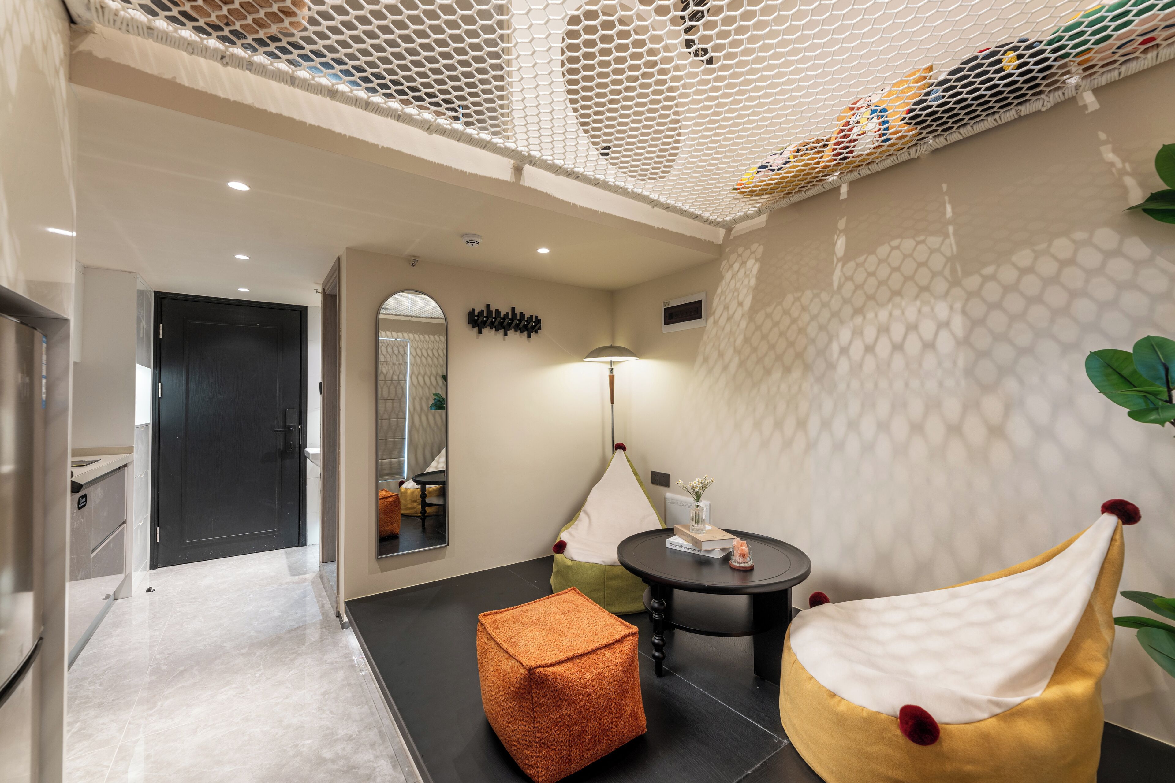 A modern interior with a black floor, a white wall with a honeycomb pattern, a round table with a lamp, and a yellow chair with red pom-poms.