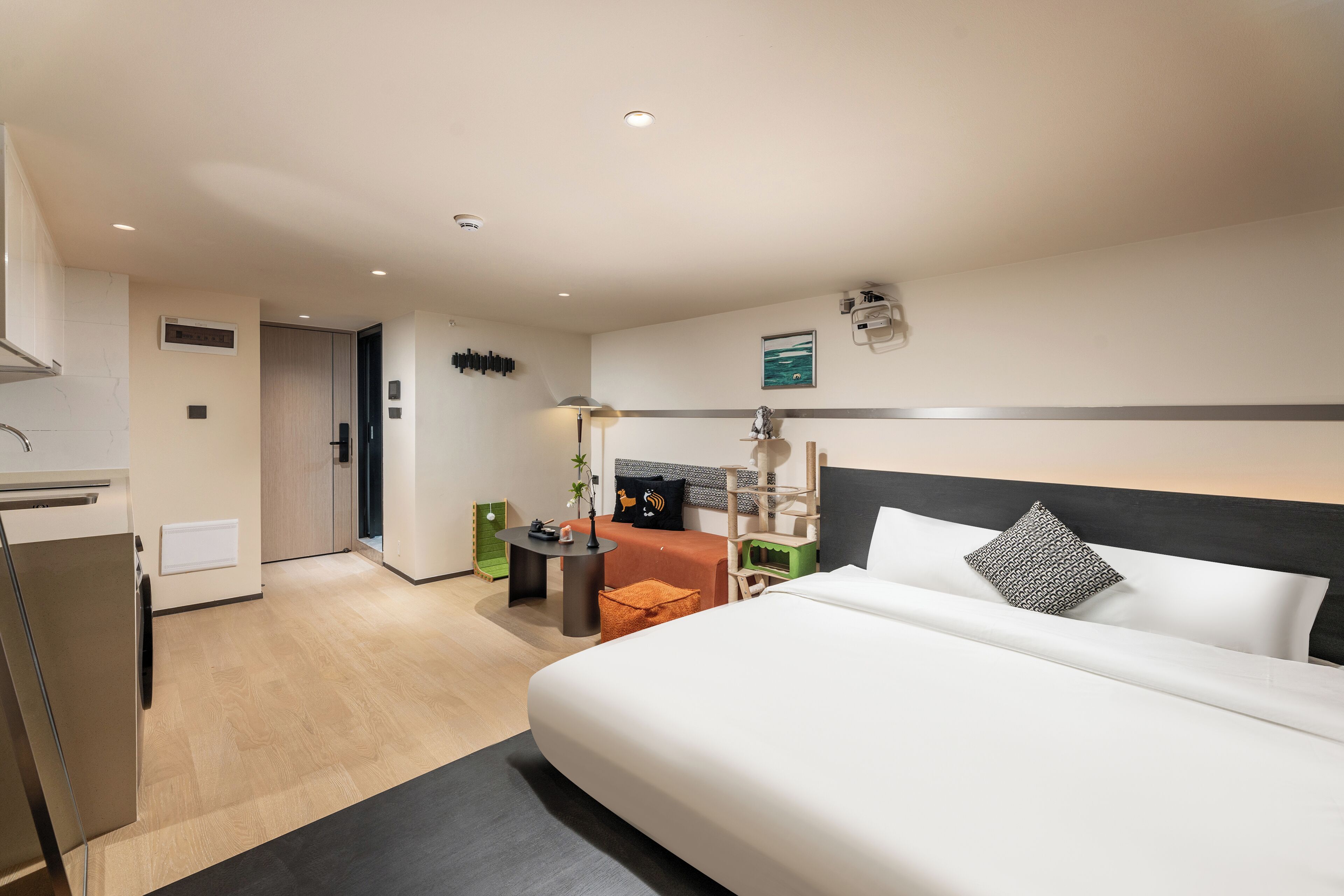 A modern hotel room with a large bed, a desk, and a chair. There is a small table with a lamp, and a green chair. The room has a wooden floor and a white wall with a picture.