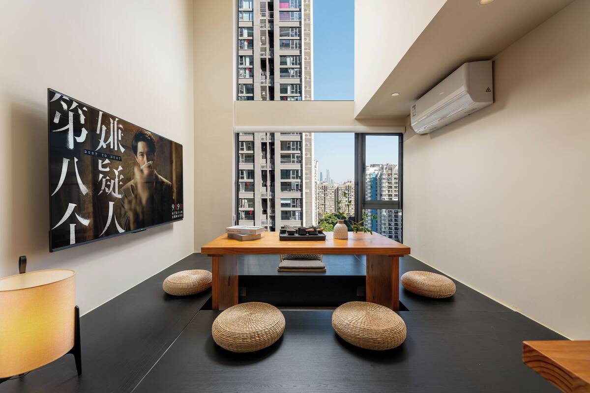 A modern interior with a wooden dining table, woven stools, a large framed artwork, and a view of high-rise buildings through large windows.