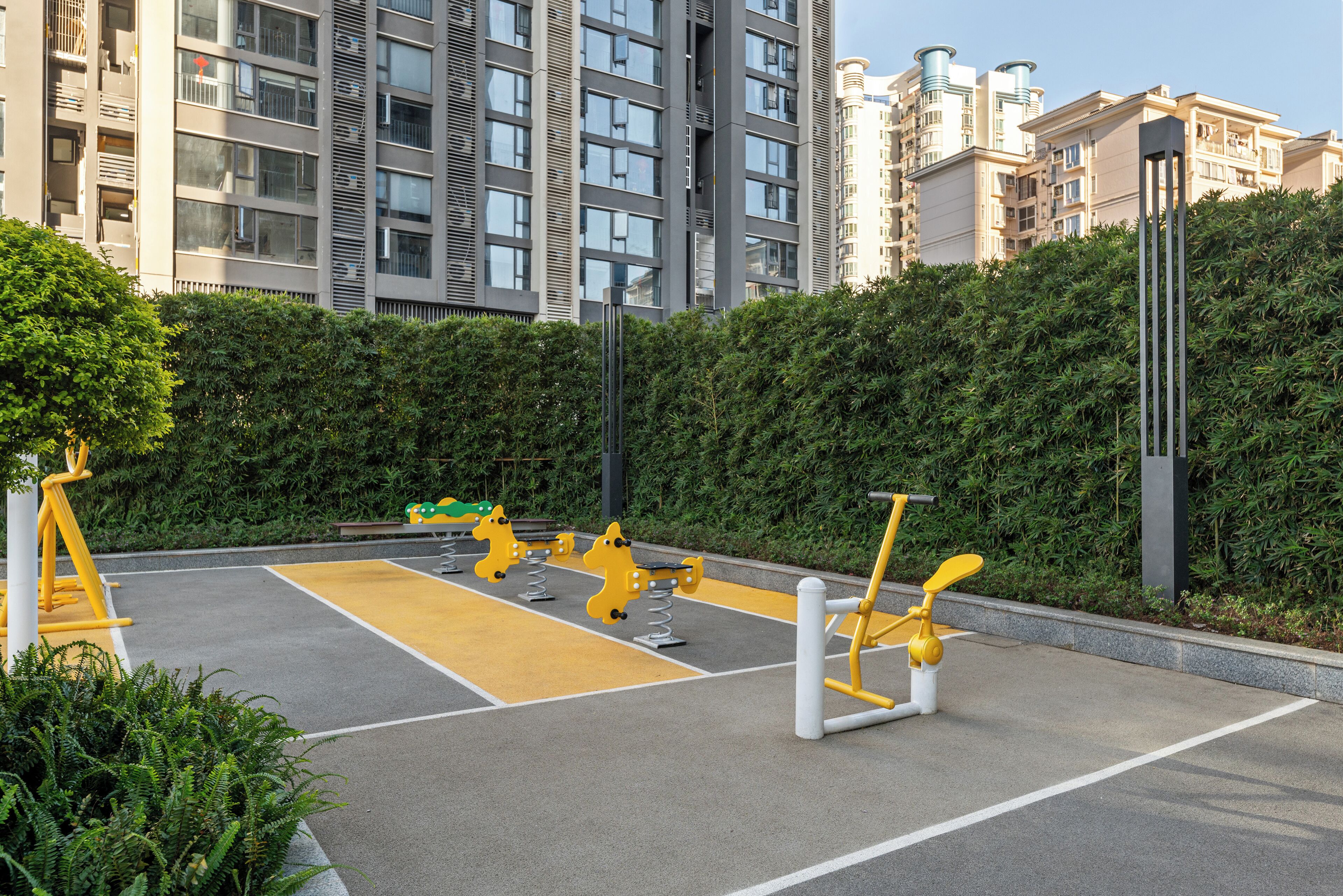 An outdoor gym area with exercise equipment, surrounded by a hedge and modern buildings in the background.