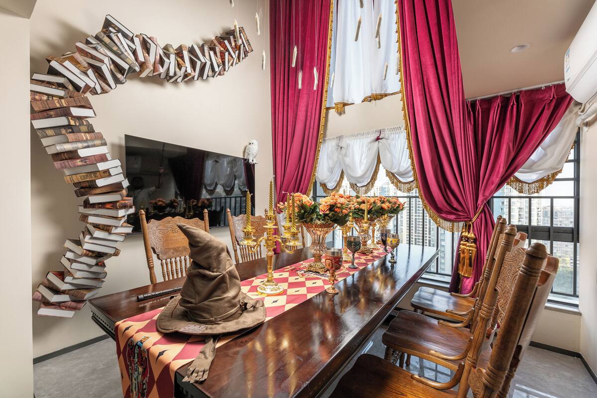 A dining area with a long table, chairs, a decorative spiral of books, a fireplace, and large windows with curtains.