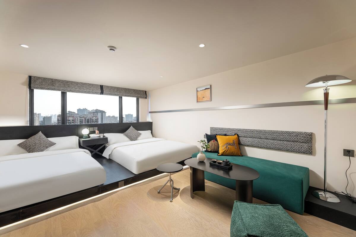 A modern hotel room with two beds, a desk, a green sofa, and a city view.