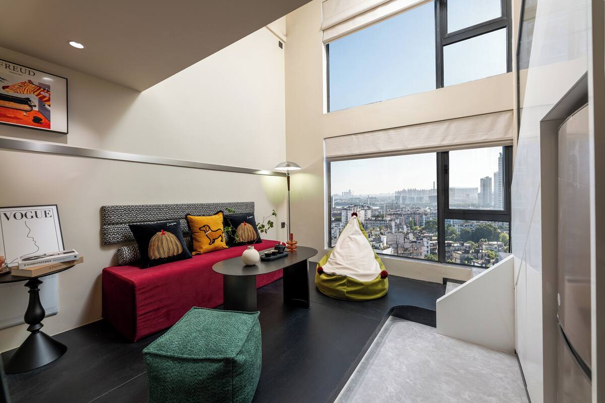 A modern living room with a red sofa, a green bean bag, a black coffee table, and a city view through large windows.