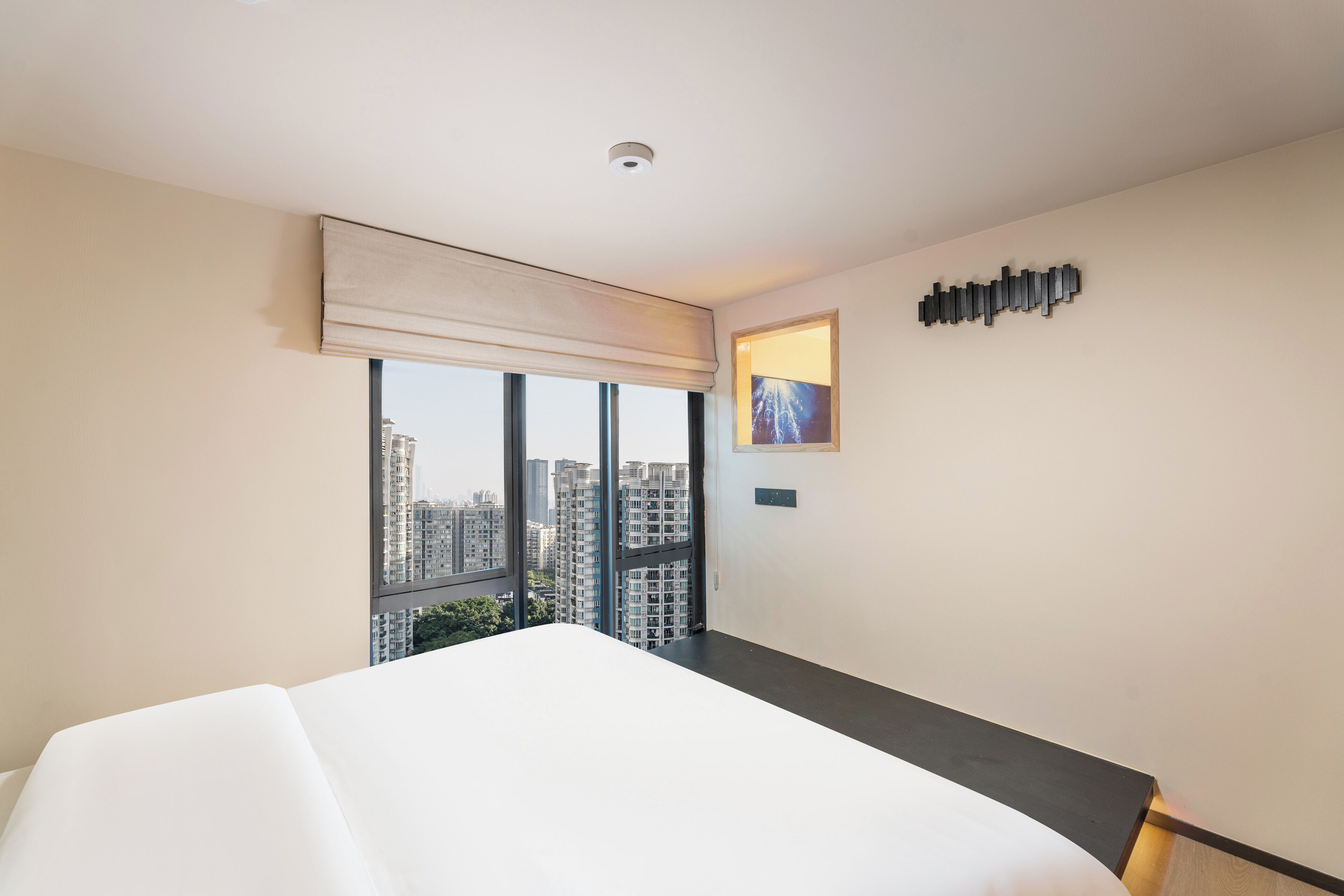A hotel room with a large bed, a window with a view of high-rise buildings, a wall-mounted artwork, and a modern light fixture.
