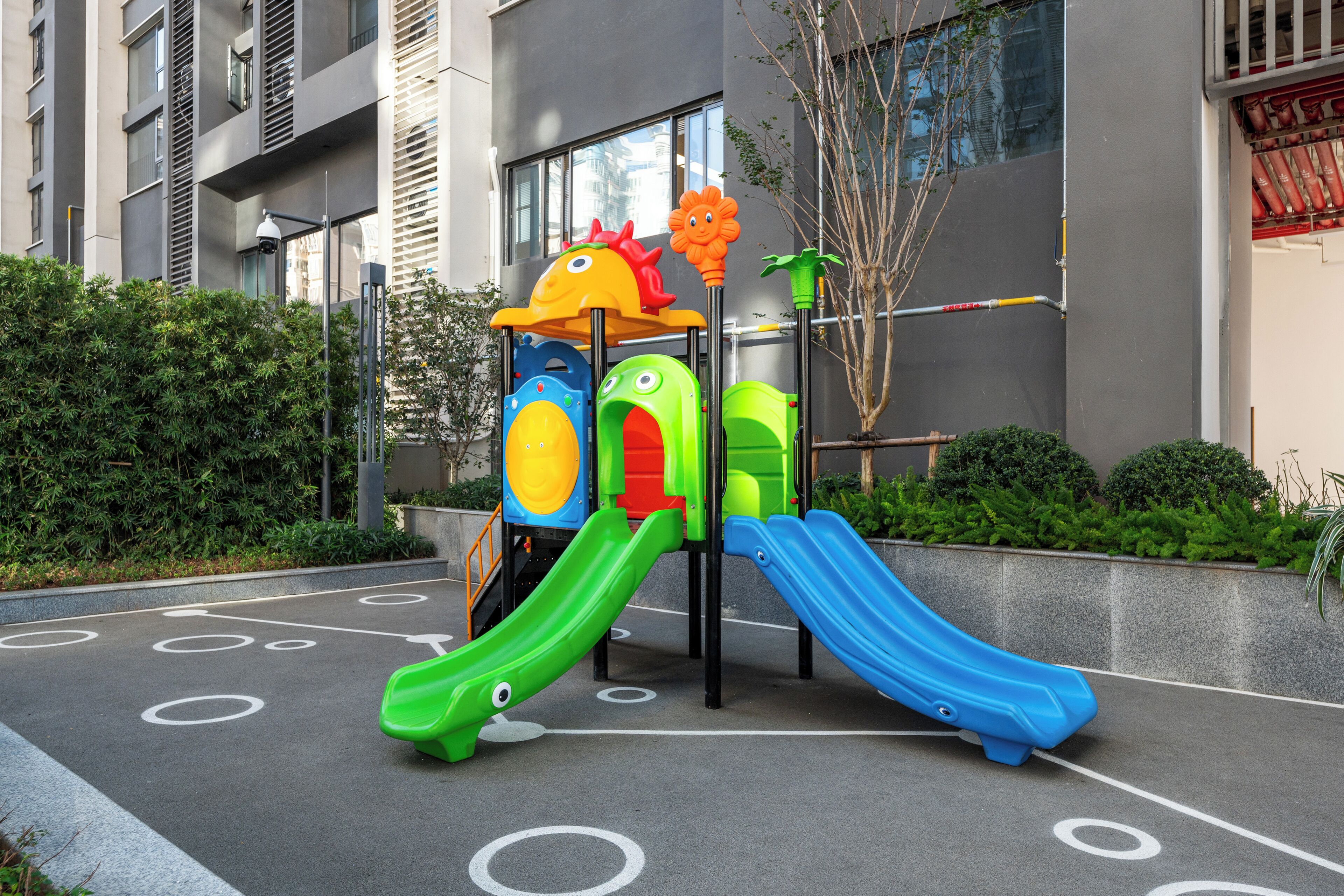 A playground with slides and a climbing structure, surrounded by a building and greenery.