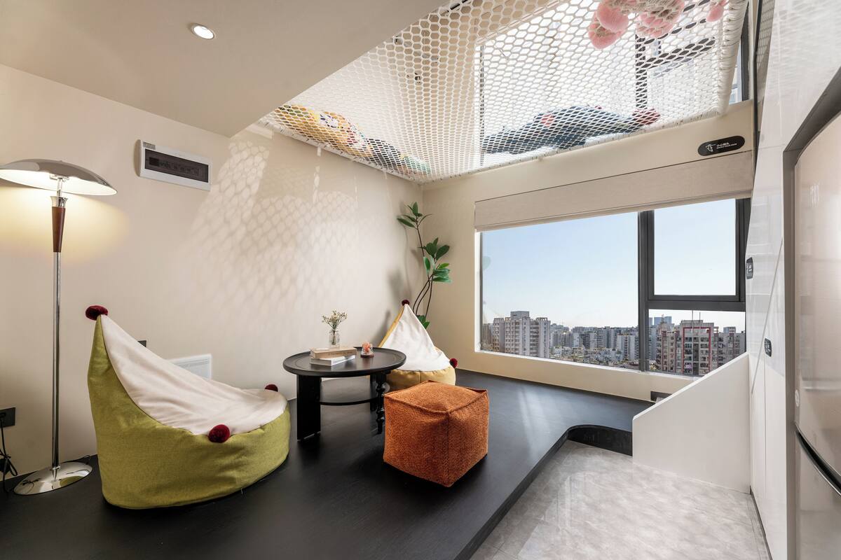 A modern room with a black table, two bean bags, a small orange ottoman, a white hanging chair, a round table, and a large window with a city view.