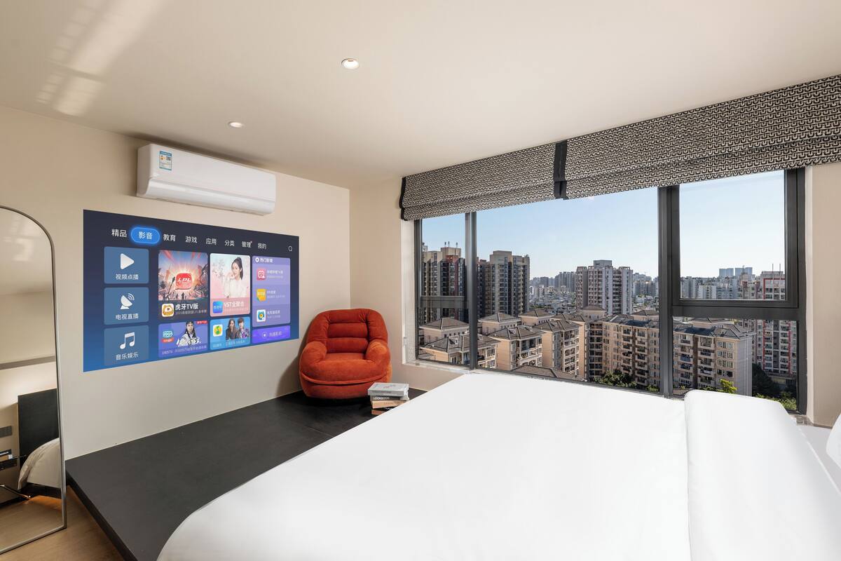A modern hotel room with a large bed, a red armchair, a mounted TV displaying various apps, and a city view through large windows.