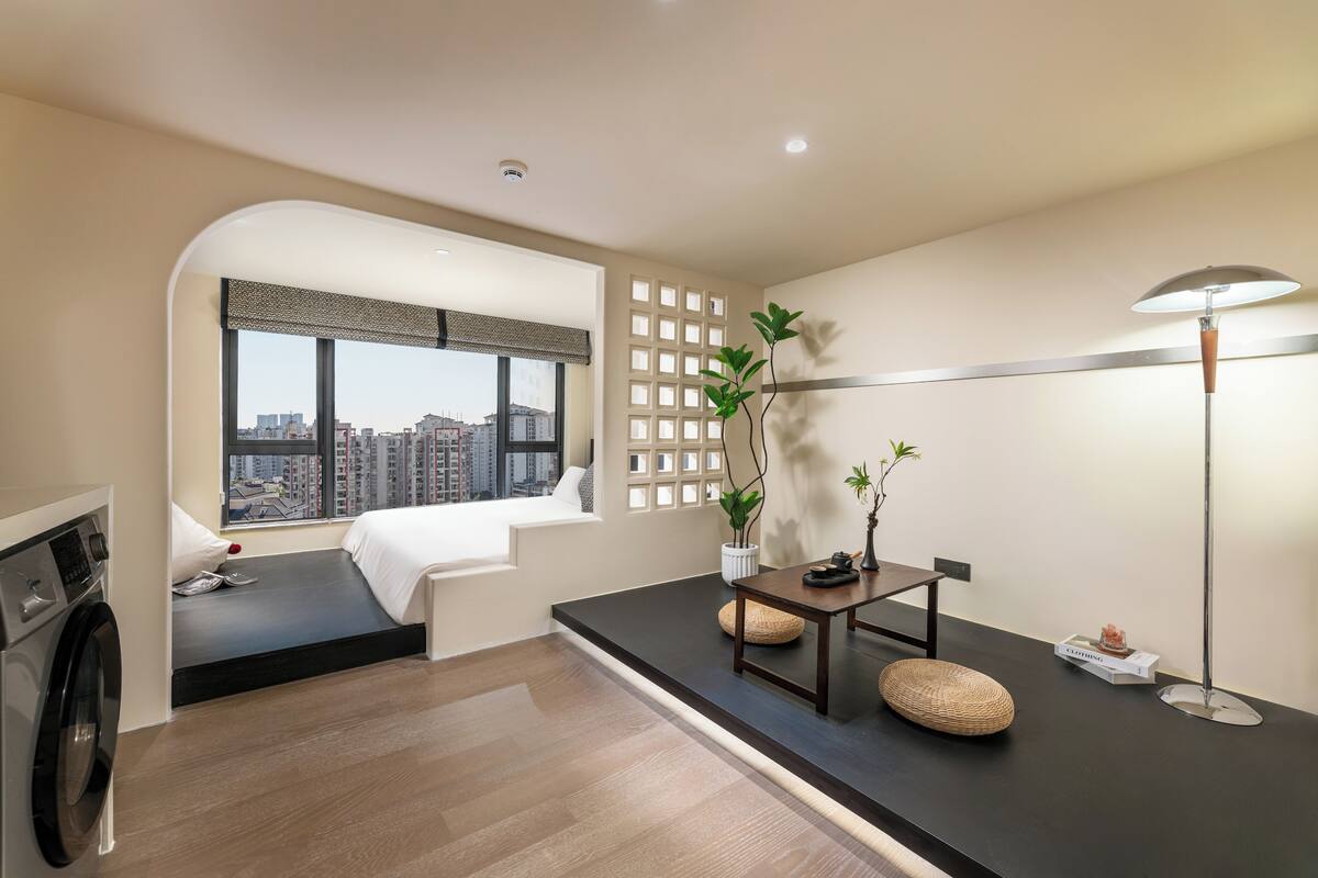 A modern hotel room with a large window, a washing machine, a small seating area, and a desk with a lamp.