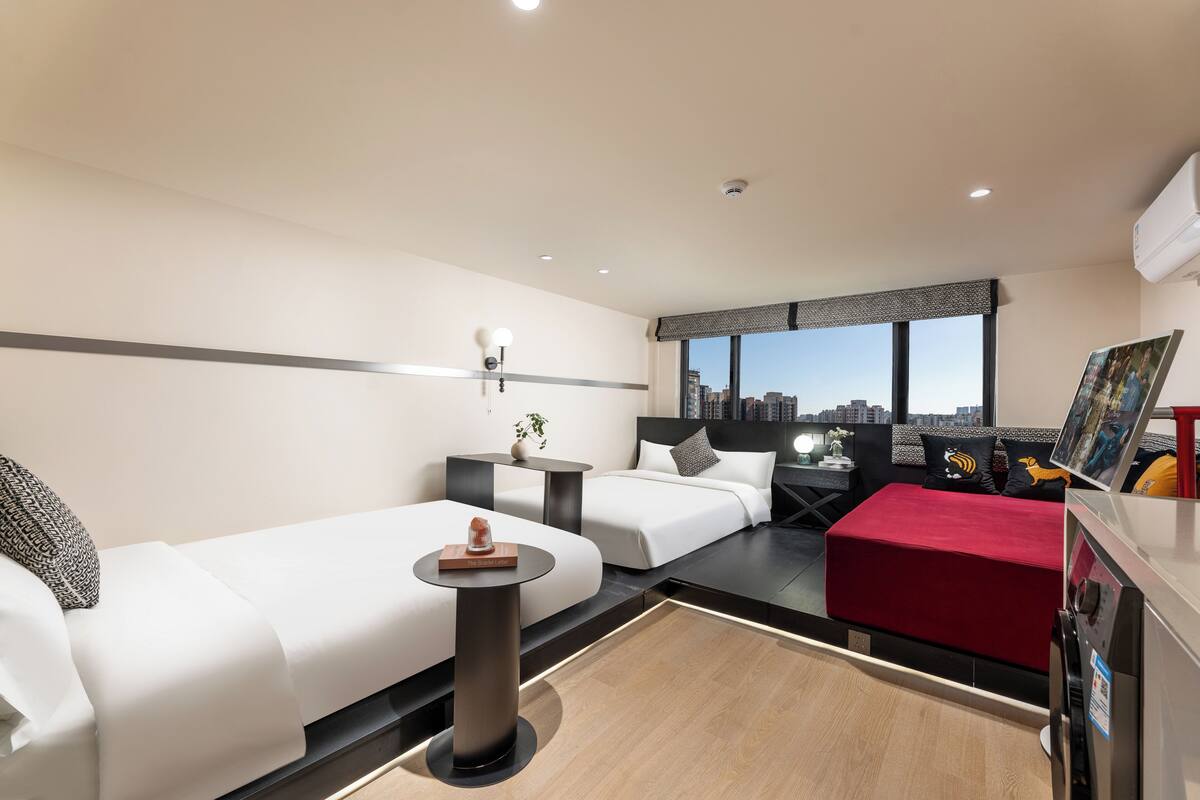 A modern hotel room with two beds, a small table, a red ottoman, and a city view.
