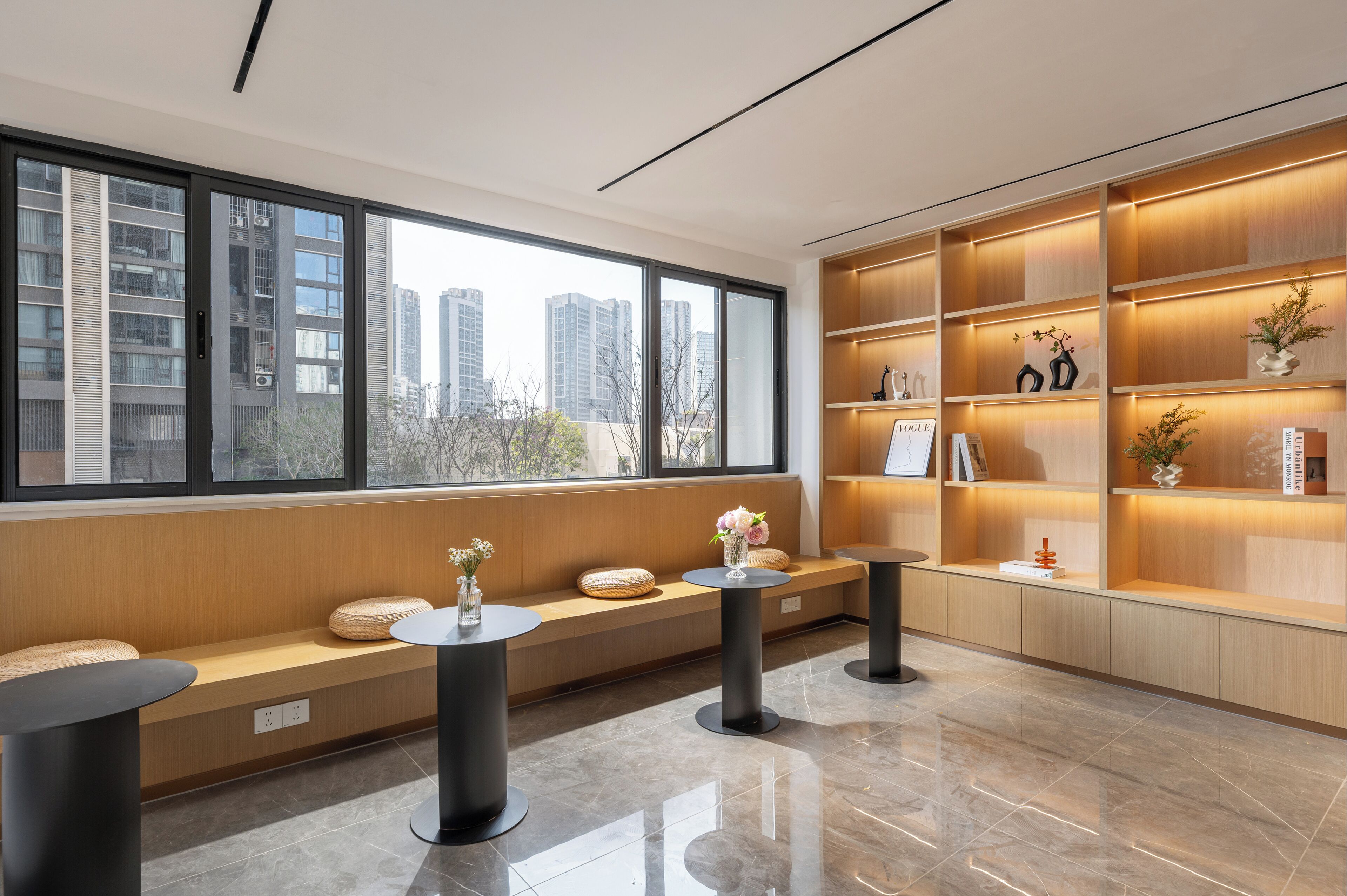 Un intérieur moderne avec une grande fenêtre, un coin salon comprenant un banc et des tables rondes, ainsi qu’une étagère en bois agrémentée d’objets décoratifs.