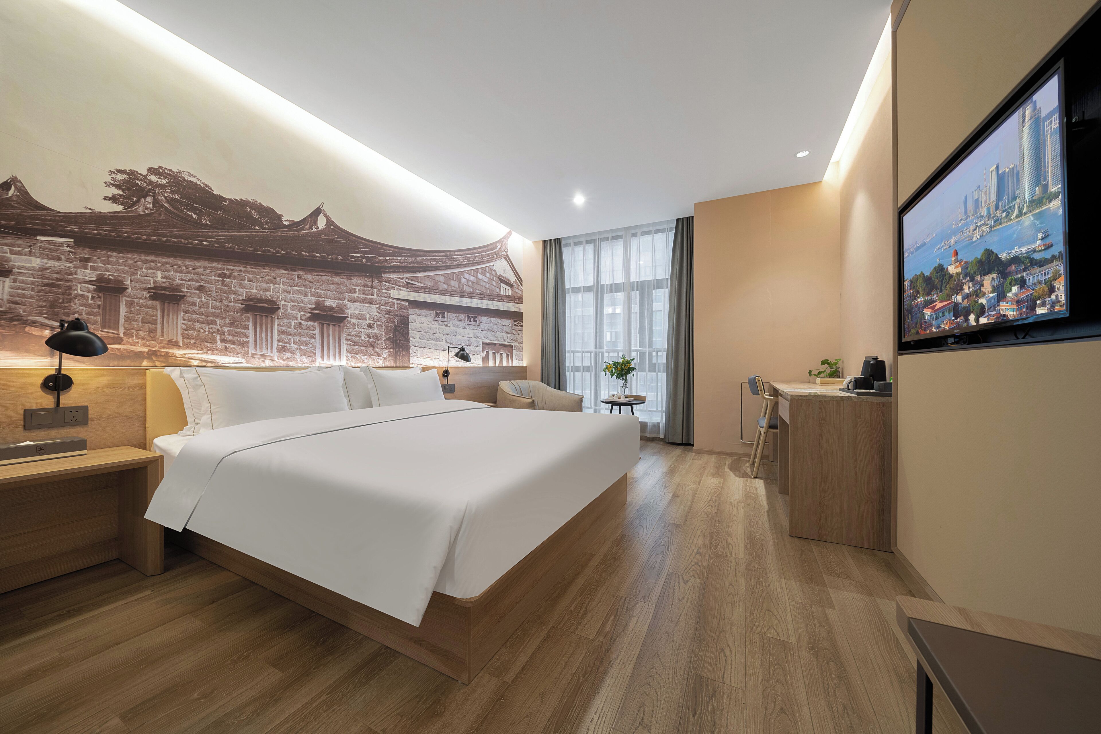 A modern hotel room with a large bed, a desk, a chair, a small table with a plant, and a TV on the wall.