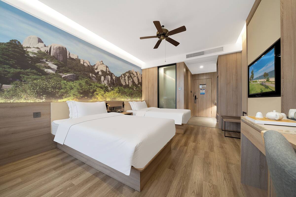 A hotel room with two beds, a ceiling fan, a TV, and a mural of a mountainous landscape.