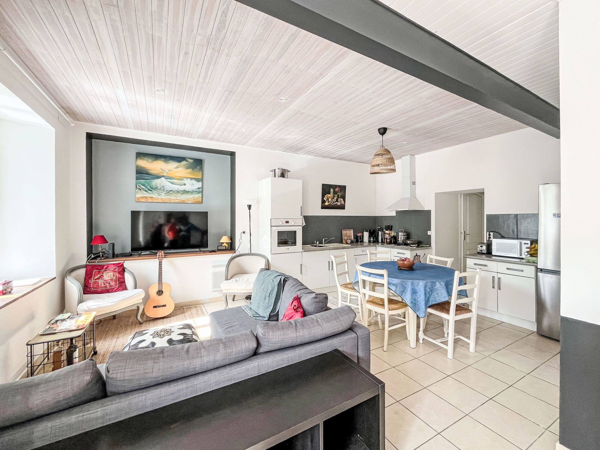 A modern living room with a grey sofa, a dining table with chairs, a kitchen area with a microwave and refrigerator, and a ceiling with wooden panels.