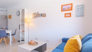 Una sala moderna con un sofá azul, una mesita de centro, una lámpara y un letrero que dice 'Welcome'. Hay una zona de comedor con sillas y una mesa, y una estantería con un reloj.