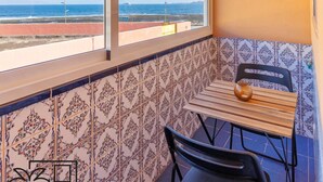 Un balcón con vistas al mar, pared de azulejos, mesa con sillas y macetas colgantes.