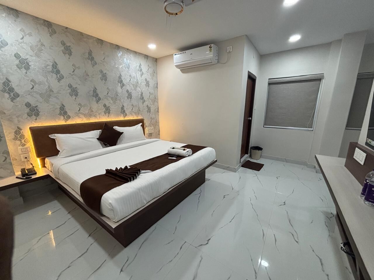 A modern hotel room with a large bed, marble flooring, a built-in headboard, an air conditioning unit, and a small bedside table.