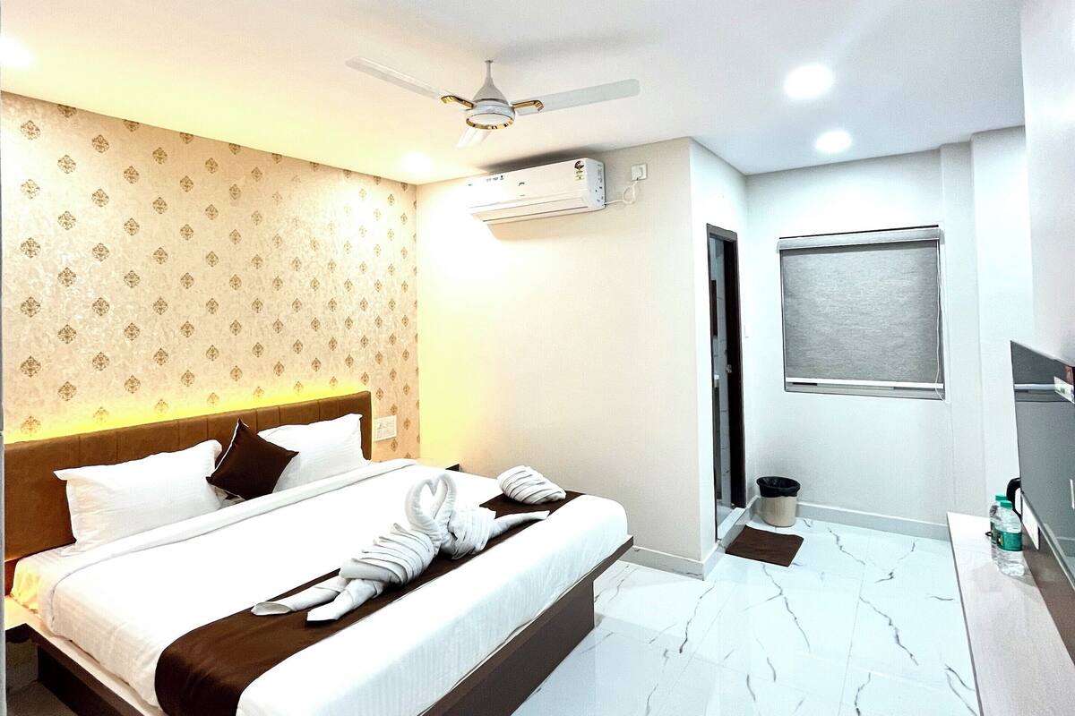 A hotel room with a large bed, a ceiling fan, an air conditioning unit, a small window, and a bathroom with a mirror and sink.