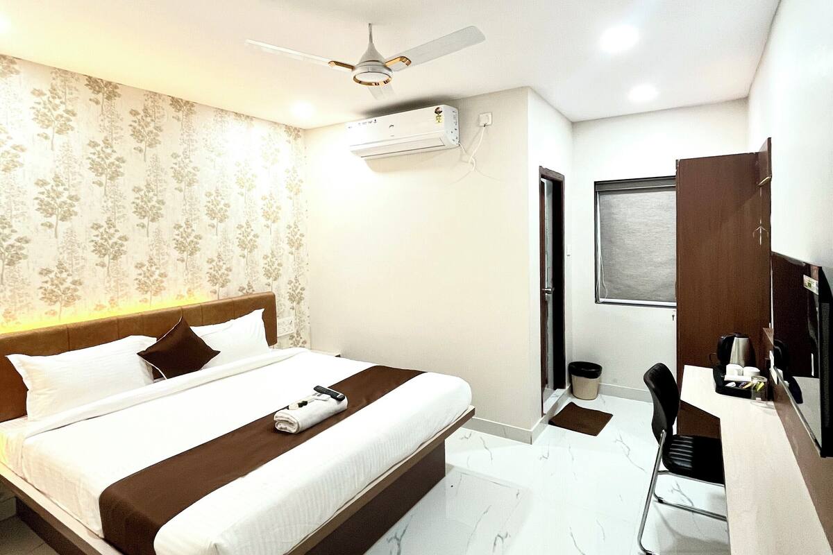 A hotel room with a bed, a desk, a chair, a ceiling fan, an air conditioning unit, and a window.
