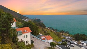 Un quartier résidentiel situé sur la côte, avec des maisons, une route asphaltée et une vue sur la mer au coucher du soleil.