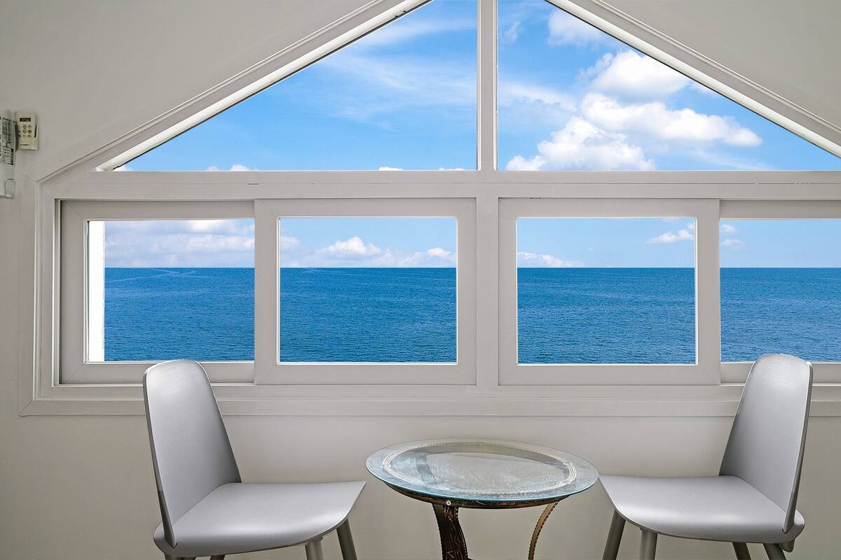 Ein Zimmer mit einem großen Fenster und Blick auf den Ozean. Ein runder Tisch mit zwei Stühlen steht darin.