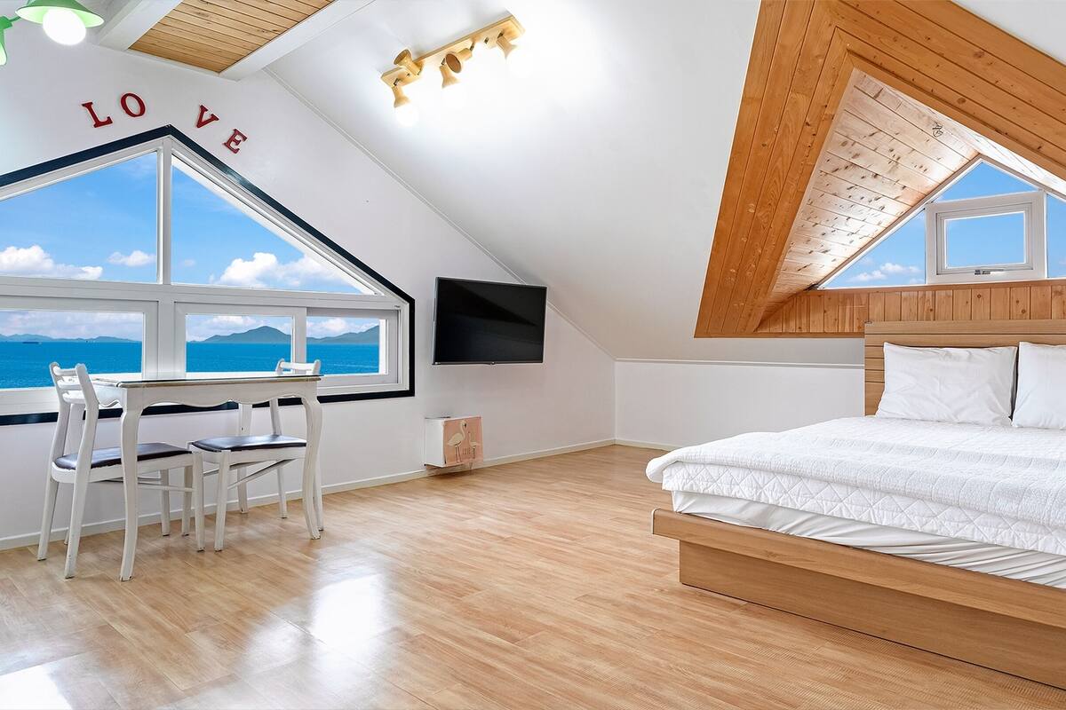 Ein Schlafzimmer mit Holzbalkendecke, einem Bett, einem Schreibtisch mit Stühlen, einem Fernseher und einem großen Fenster mit Meerblick.