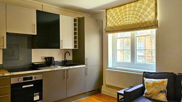 Une cuisine compacte dotée d’appareils électroménagers modernes, une fenêtre agrémentée de rideaux à motifs et un canapé orné d’un coussin décoratif.