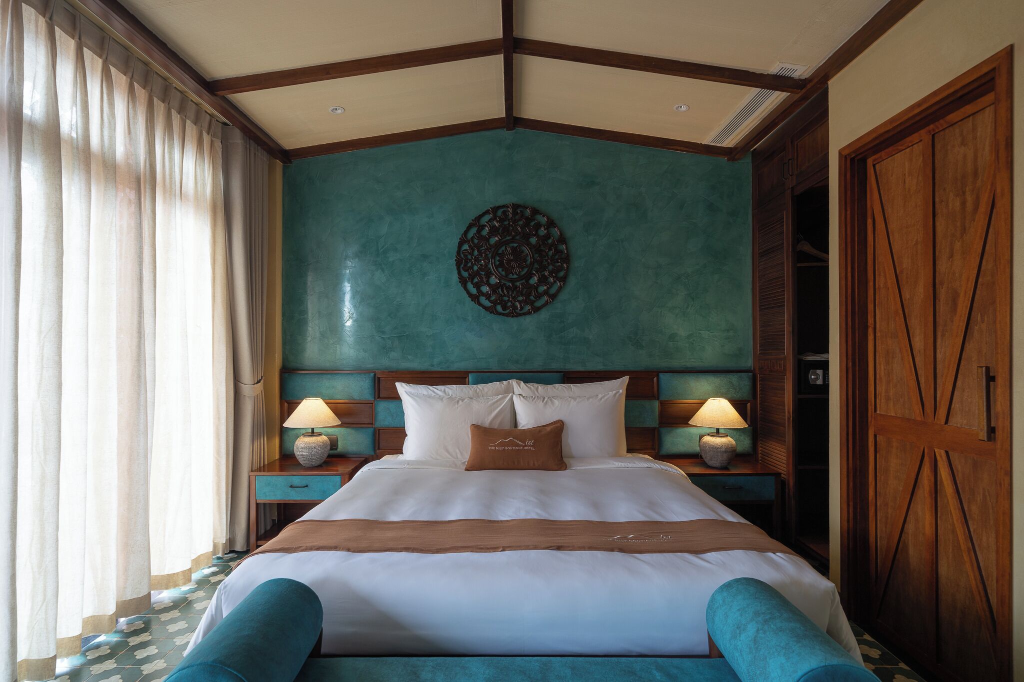 A hotel room with a large bed, two bedside tables with lamps, a wooden wardrobe, and a decorative wall piece.