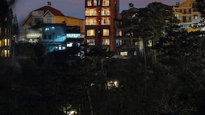 Un edificio de varios pisos con ventanas iluminadas al anochecer, rodeado de árboles y otros edificios.