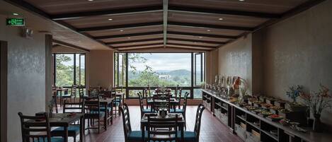 A restaurant with wooden tables and chairs, a long counter with food displays, and large windows offering a view of greenery and hills.