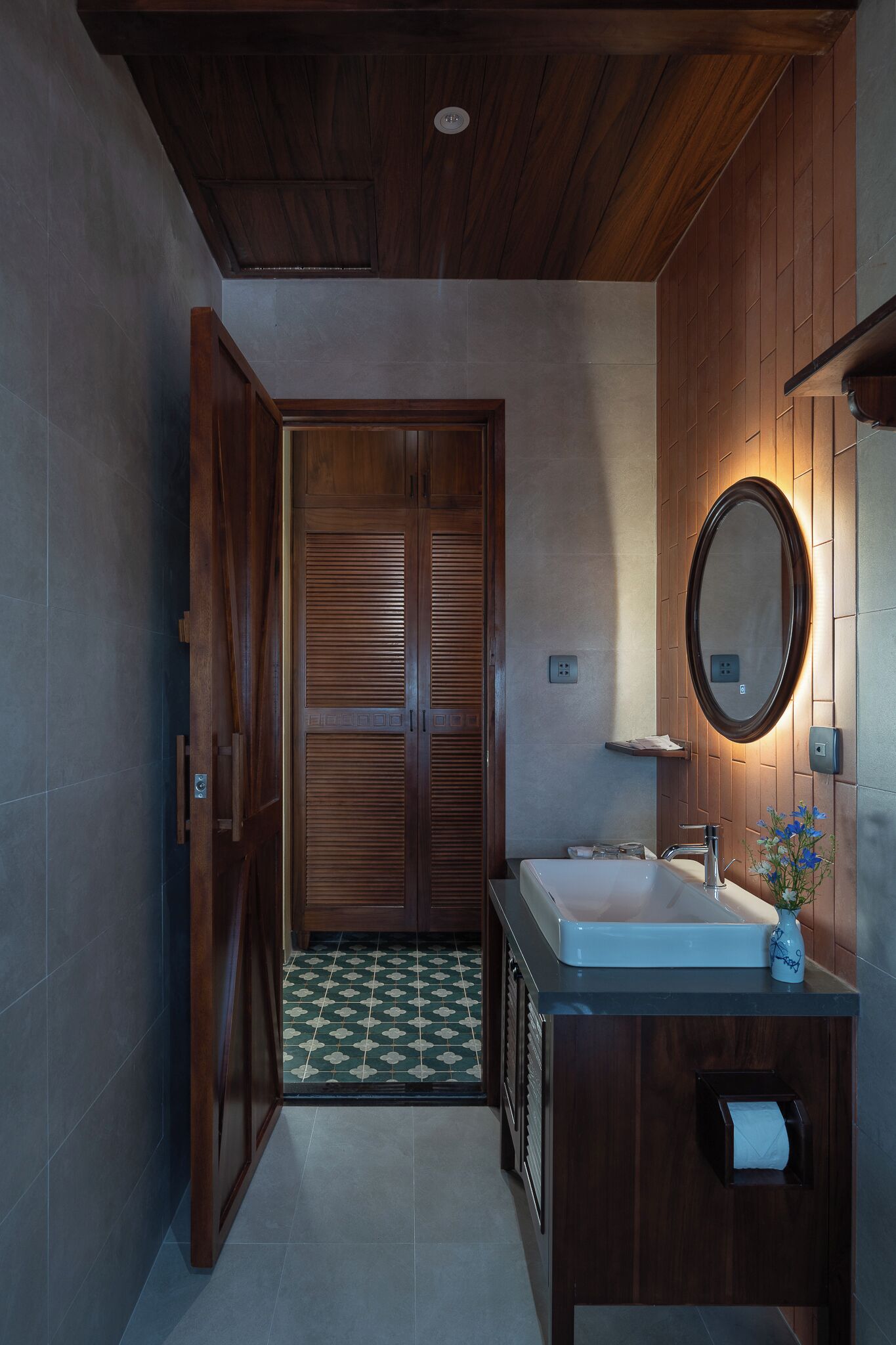 A bathroom with a wooden door, a sink, a mirror, and a tiled floor.
