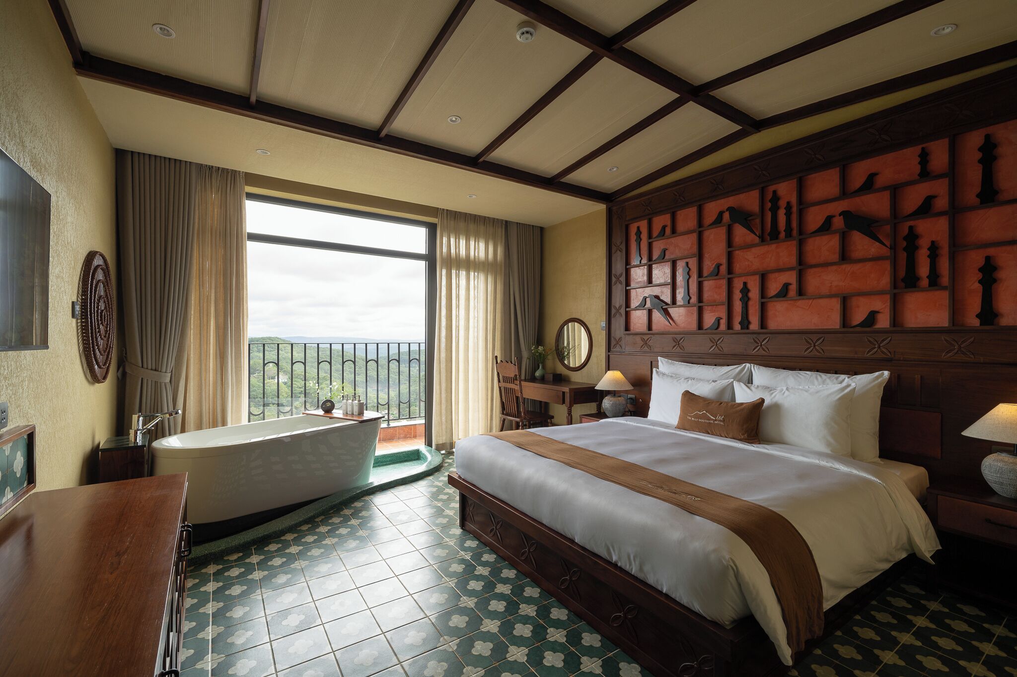 A hotel room with a large bed, a bathtub, a wooden headboard with carvings, a view of the outdoors, and a tiled floor.