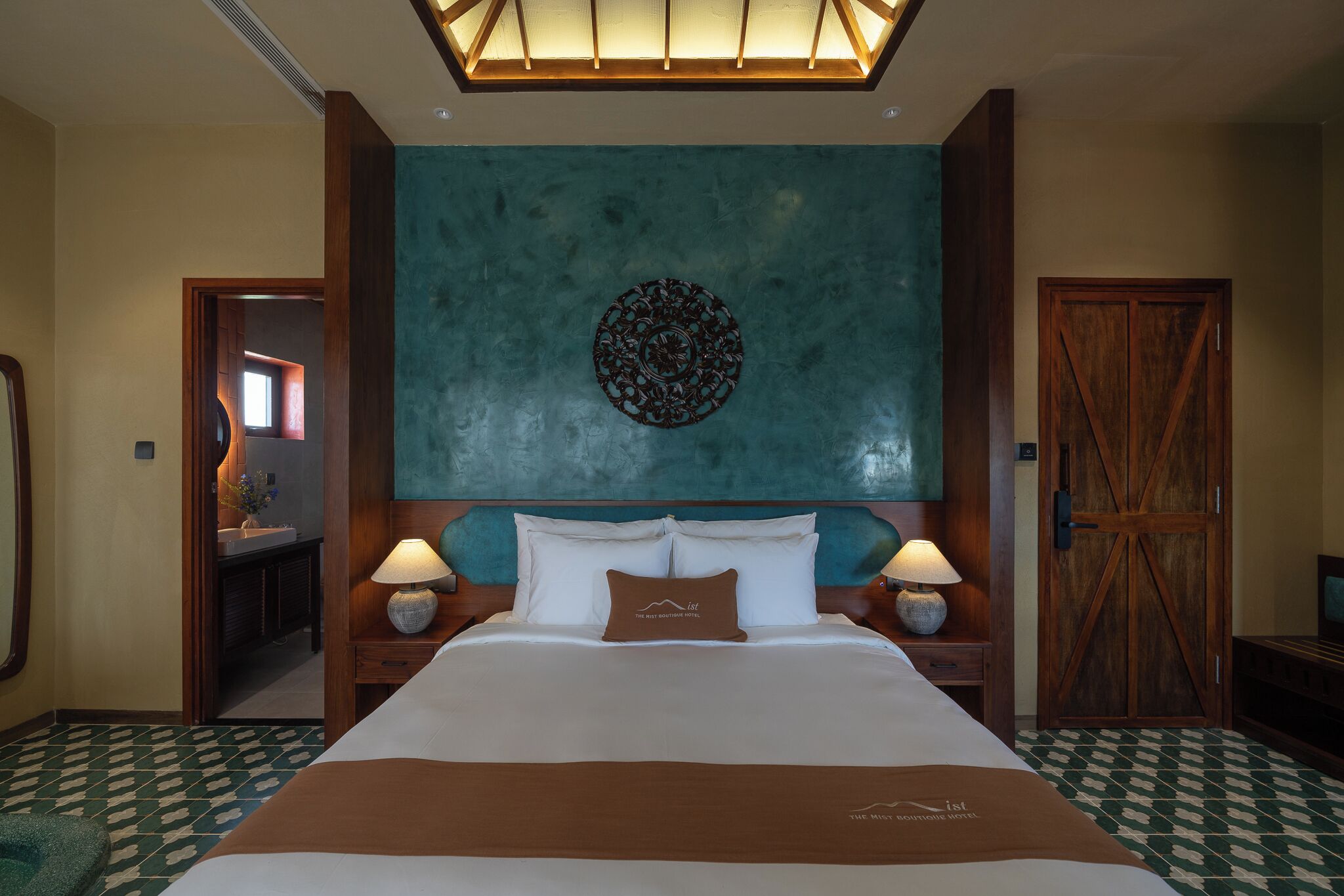 Habitación de hotel con una cama grande, dos mesitas de noche con lámparas, una puerta de madera y un adorno decorativo sobre la cama.