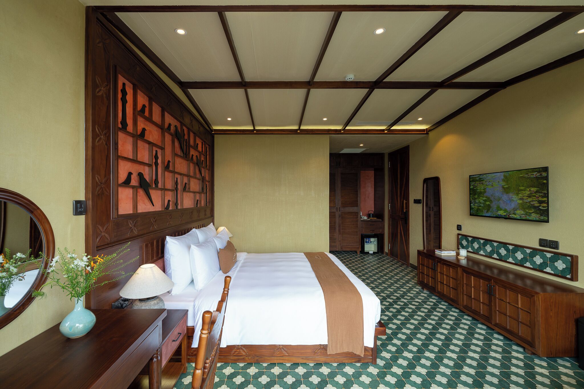 A hotel room with a large bed, a wooden headboard, a bedside table with a lamp, a mirror, and a vase with flowers.