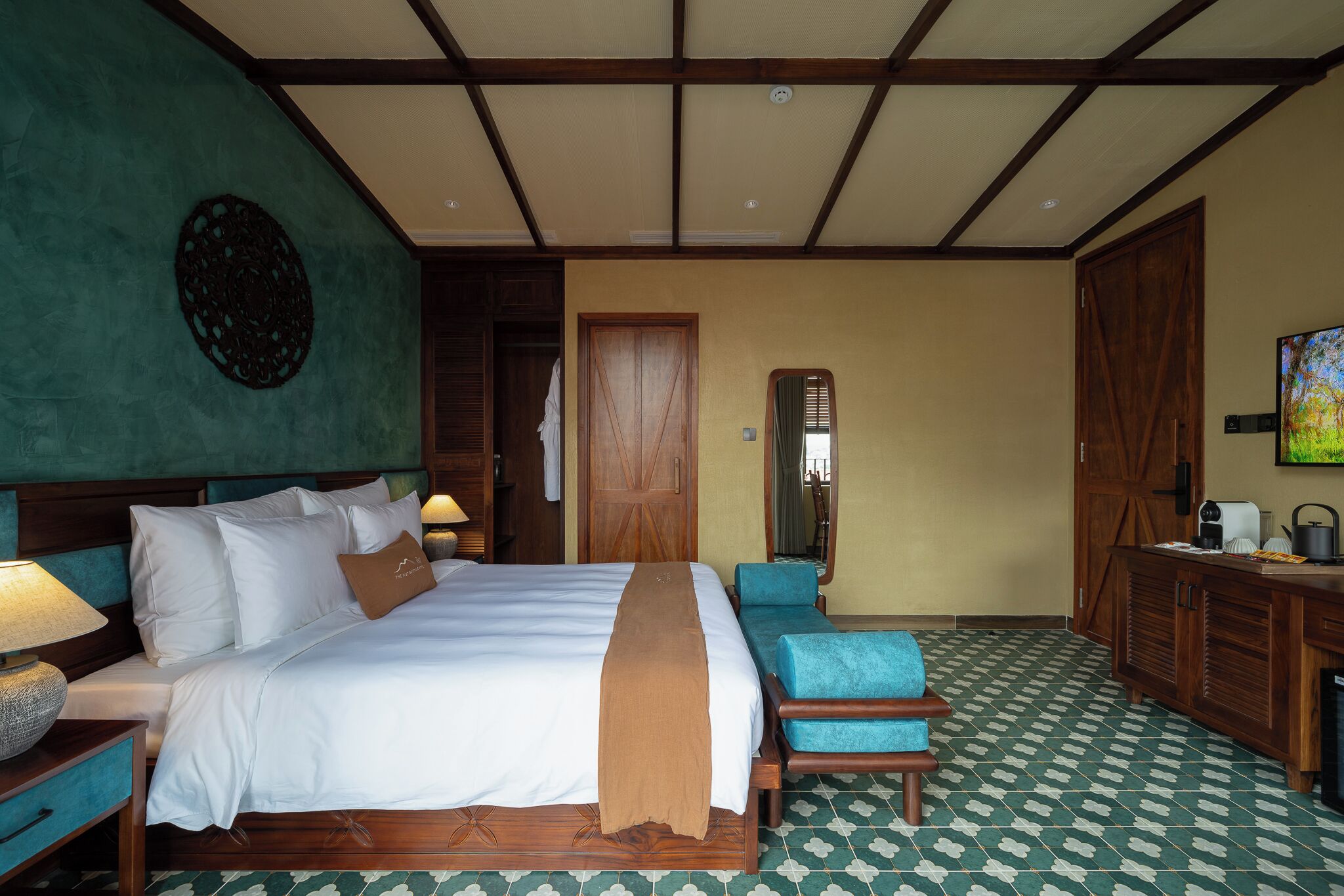 Una habitación de hotel con una cama grande, un cabecero de madera, un sillón azul, un aparador de madera y un suelo con estampado verde.