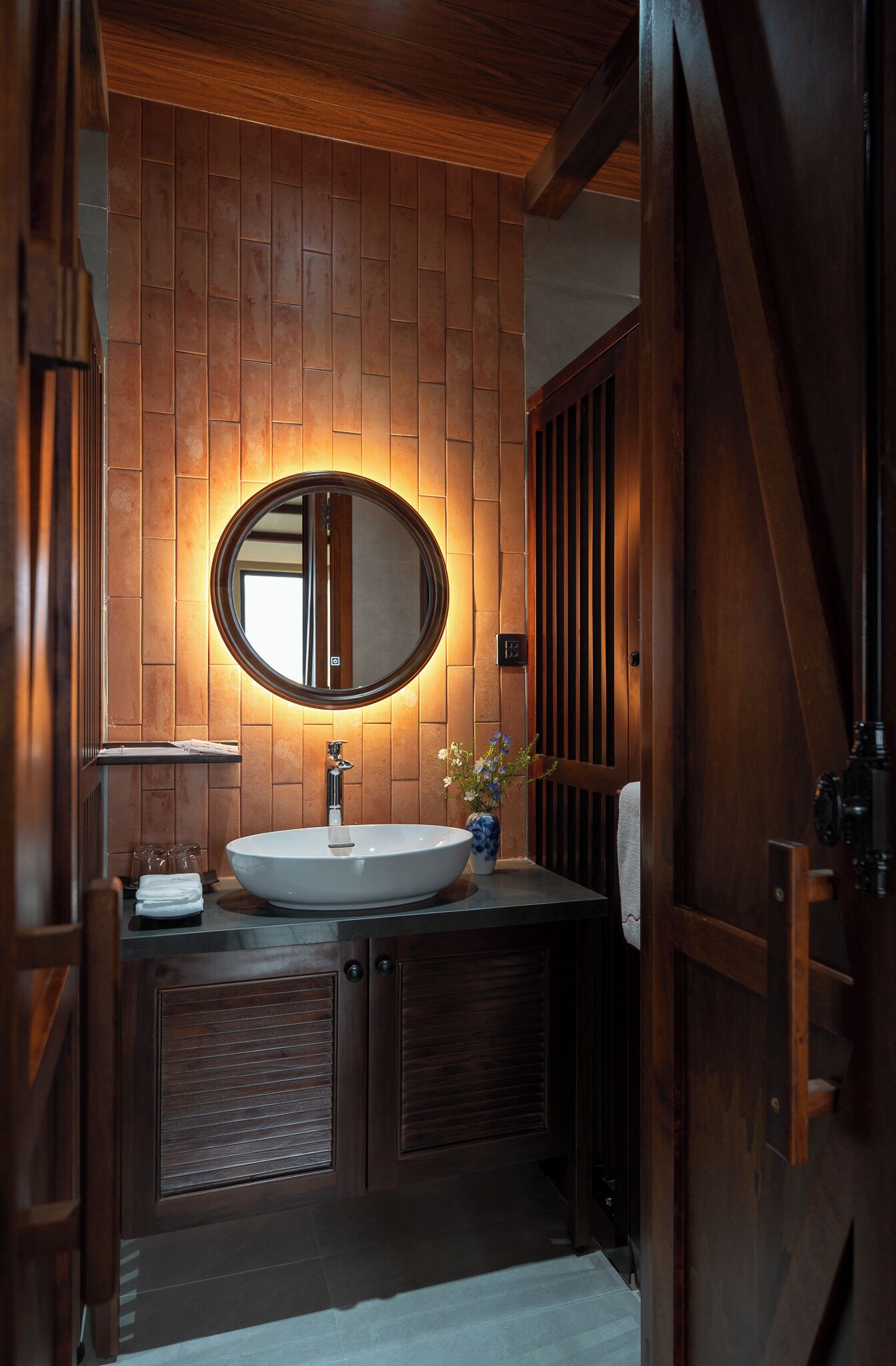 A bathroom with a wooden panel wall, a round mirror, a white sink, and a vase with flowers.